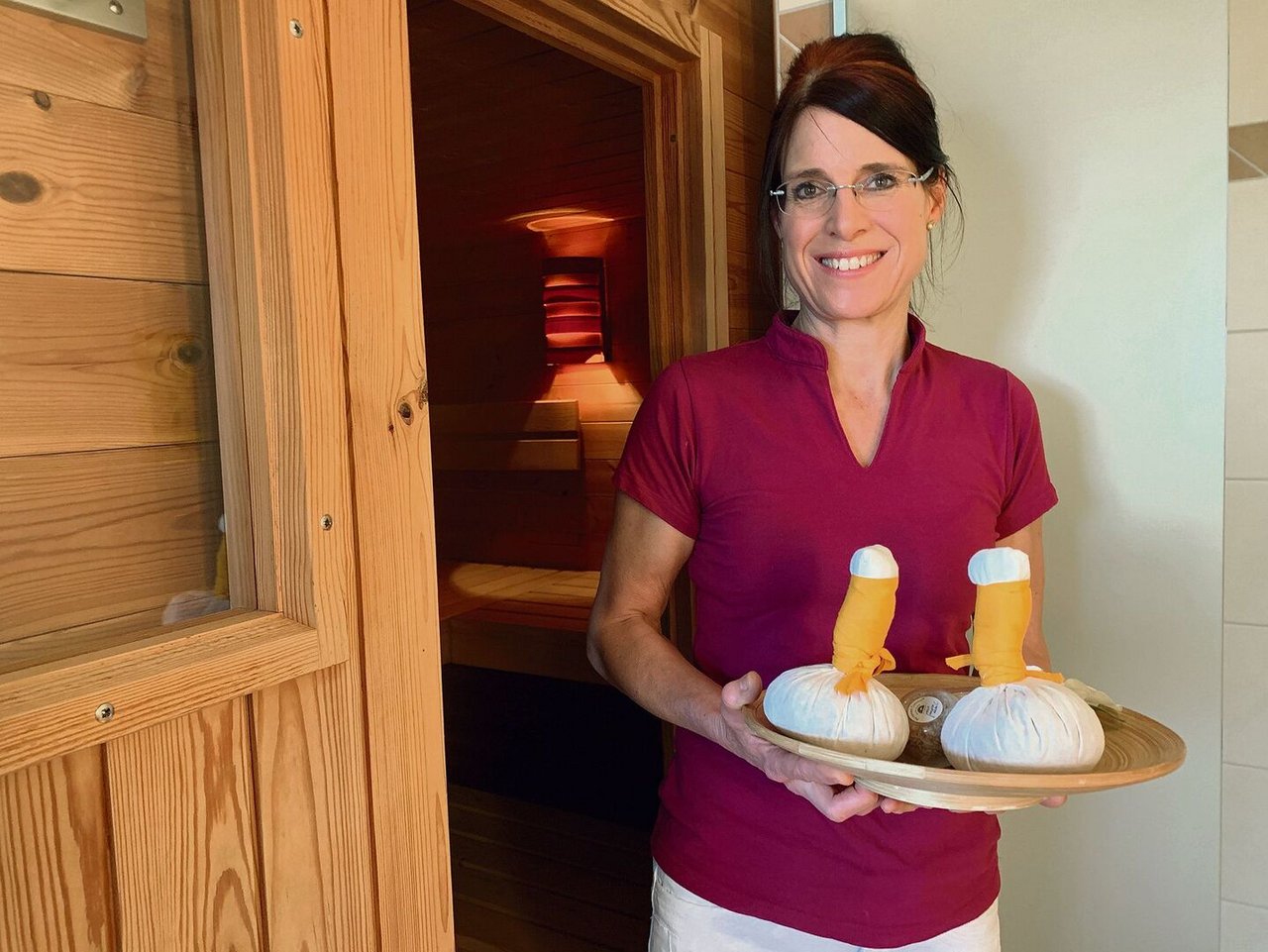 Fabienne Toppler posiert vor der Sauna, auf dem Tablett liegen Kräuter- und Blütenstempel für ihre Wellnessbehandlungen parat.