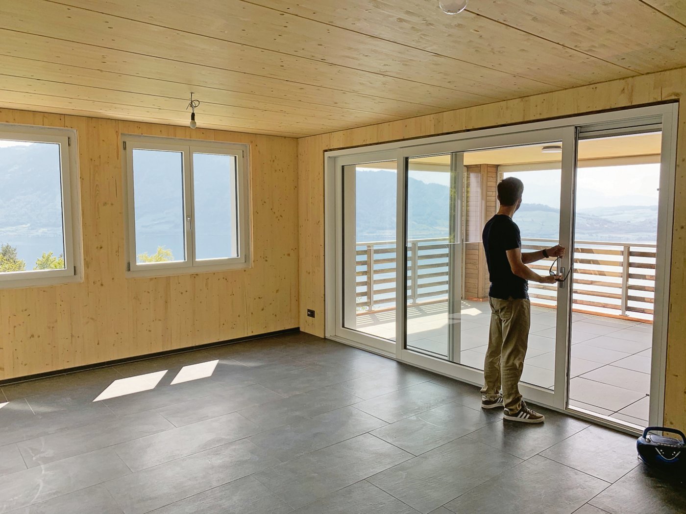 Blick in den Wohnraum mit grosszügigem Balkon und prächtiger Aussicht. Der Bodenaufbau ist aus Holz, darüber wurden in diesem Raum Steinplatten verlegt.