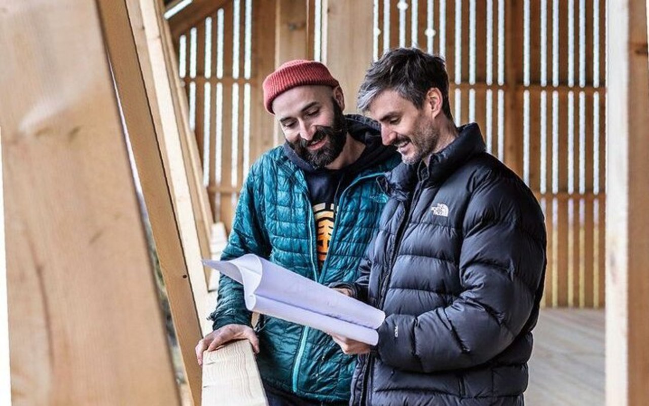 Lionel Richard (links) und Michael Haenni stehen am Holzgeländer und schauen gemeinsam in die Baupläne.