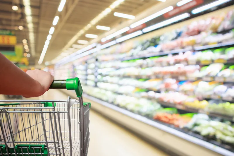 Eine Hand schiebt einen leeren Einkaufswagen durch die Regale eines Supermarkts.